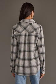 A woman with long brown hair, wearing gold hoop earrings, and dressed in the Panache Apparel Collection’s Light Gray, Black & Sage Plaid Button Up and blue jeans, stands facing away from the camera against a plain gray background.