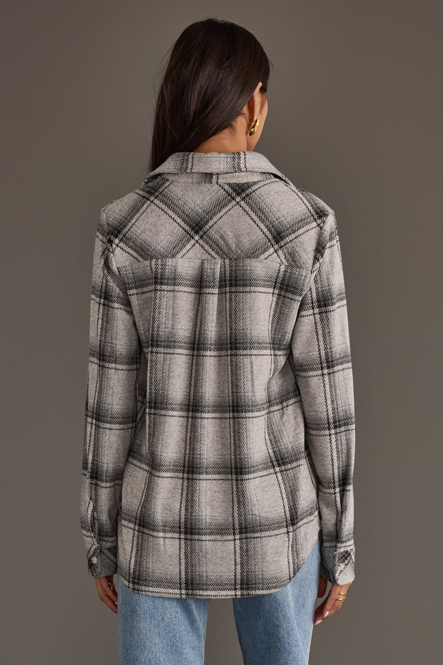 A woman with long brown hair, wearing gold hoop earrings, and dressed in the Panache Apparel Collection’s Light Gray, Black & Sage Plaid Button Up and blue jeans, stands facing away from the camera against a plain gray background.
