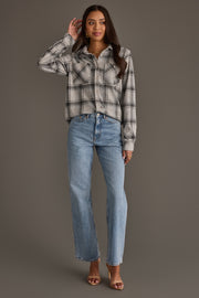 A woman stands against a gray background, wearing the Panache Apparel Collection Light Gray, Black & Sage Plaid Button Up paired with light blue jeans and brown heeled sandals. She has long dark hair and touches her head with one hand while looking at the camera.