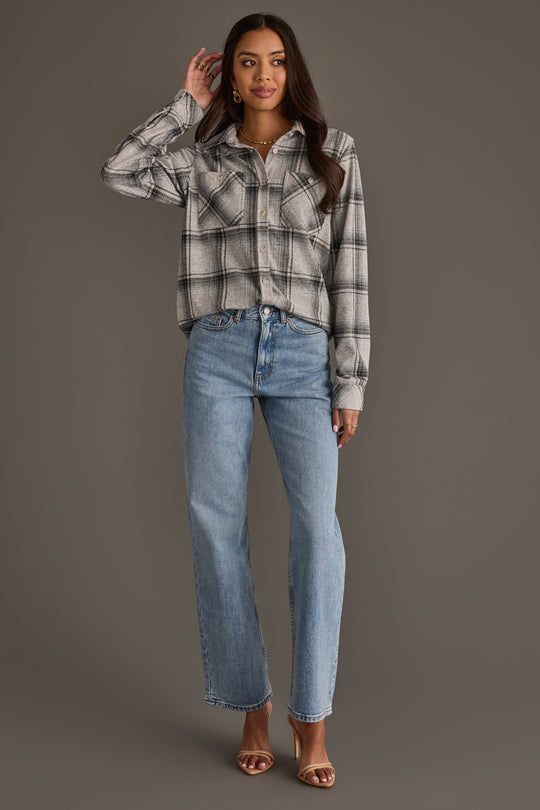 A woman stands against a gray background, wearing the Panache Apparel Collection Light Gray, Black & Sage Plaid Button Up paired with light blue jeans and brown heeled sandals. She has long dark hair and touches her head with one hand while looking at the camera.