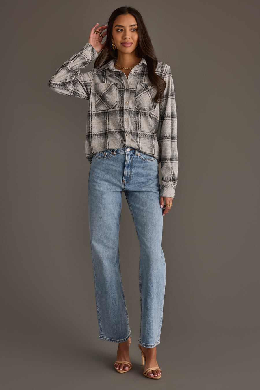 A woman stands against a gray background, wearing the Panache Apparel Collection Light Gray, Black & Sage Plaid Button Up paired with light blue jeans and brown heeled sandals. She has long dark hair and touches her head with one hand while looking at the camera.