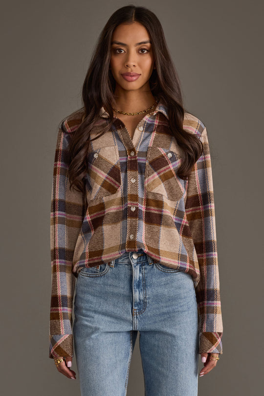 A woman with long brown hair wears the Panache Apparel Collection’s Dark Brown, Blue & Pink Plaid Button Up shirt and light blue jeans, standing against a plain gray background.