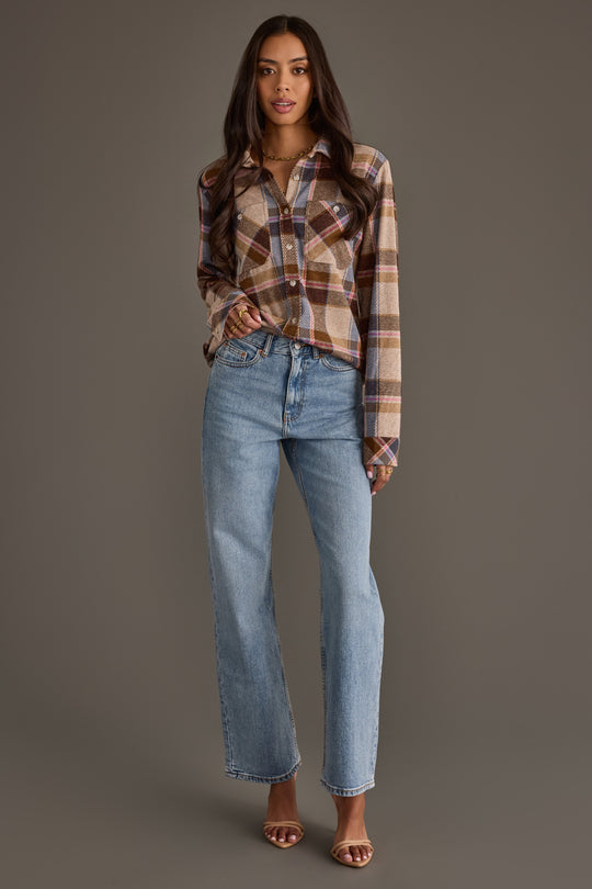 A woman stands against a plain background, wearing the Panache Apparel Collection’s Dark Brown, Blue & Pink Plaid Button Up paired with light blue high-waisted jeans and open-toed brown heels. She has long dark hair and a relaxed expression.