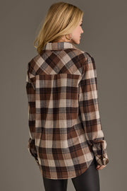 A woman with blonde hair, dressed in the Panache Apparel Collection’s Brown, Black & Cream Plaid Button Up and black pants, stands facing away from the camera against a neutral background.