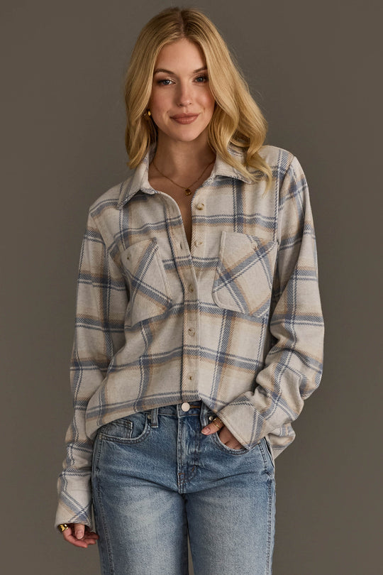A person with long blonde hair wears the Cream, Dusty Blue & Tan Plaid Button Up from Panache Apparel Collection and light blue jeans, standing against a plain gray background and smiling with one hand in a pocket.