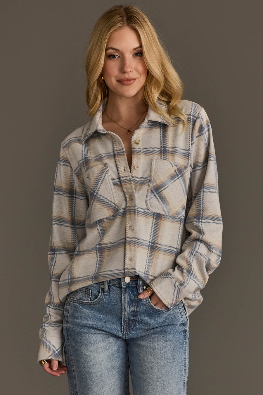 A person with long blonde hair wears the Cream, Dusty Blue & Tan Plaid Button Up from Panache Apparel Collection and light blue jeans, standing against a plain gray background and smiling with one hand in a pocket.