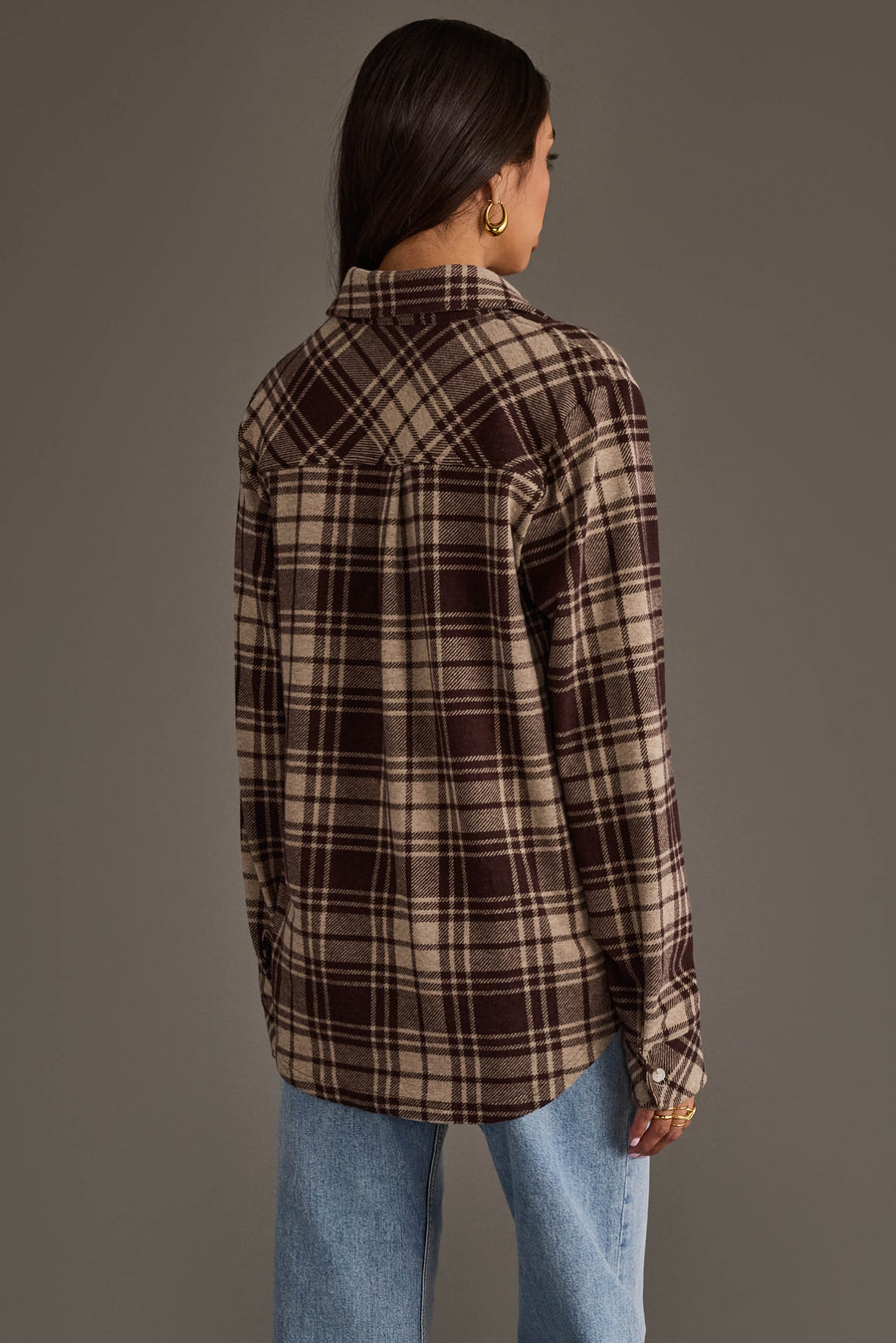 A person with long dark hair is seen from behind, standing against a plain background and wearing the Panache Apparel Collection’s Chocolate Brown & Tan Plaid Button Up along with light blue jeans.