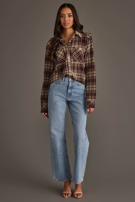A woman stands against a gray background wearing the Panache Apparel Collection Chocolate Brown & Tan Plaid Button Up, paired with light blue high-waisted jeans and open-toed heels.