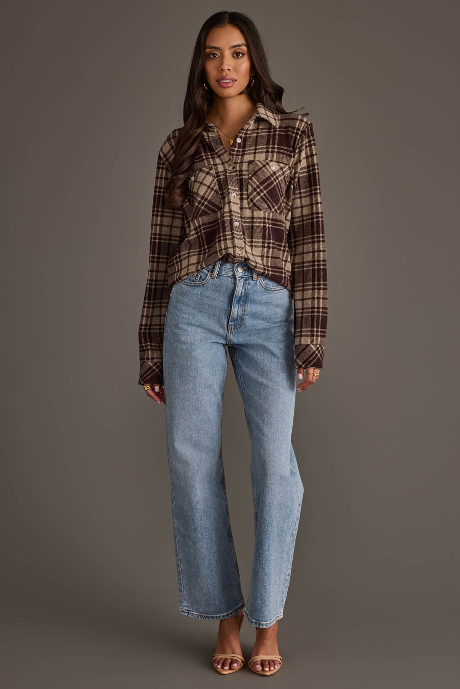 A woman stands against a gray background wearing the Panache Apparel Collection Chocolate Brown & Tan Plaid Button Up, paired with light blue high-waisted jeans and open-toed heels.