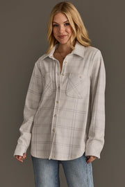 A woman with blonde hair wears the Panache Apparel Collection’s White & Light Gray Plaid Button Up, featuring two chest pockets, paired with blue jeans. She stands in front of a plain gray background and smiles slightly.