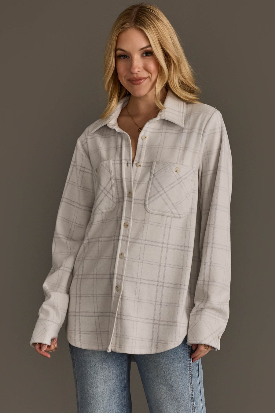 A woman with blonde hair wears the Panache Apparel Collection’s White & Light Gray Plaid Button Up, featuring two chest pockets, paired with blue jeans. She stands in front of a plain gray background and smiles slightly.