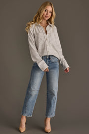A woman with long blonde hair, dressed in the Panache Apparel Collection’s White & Light Gray Plaid Button Up and blue jeans, stands against a plain brown background. She is smiling softly with one hand in her pocket.