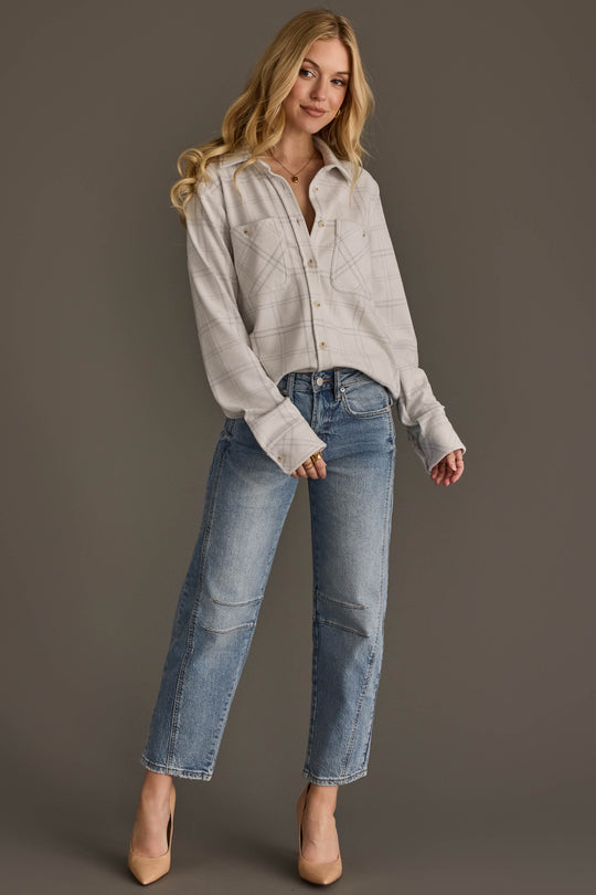 A woman with long blonde hair, dressed in the Panache Apparel Collection’s White & Light Gray Plaid Button Up and blue jeans, stands against a plain brown background. She is smiling softly with one hand in her pocket.