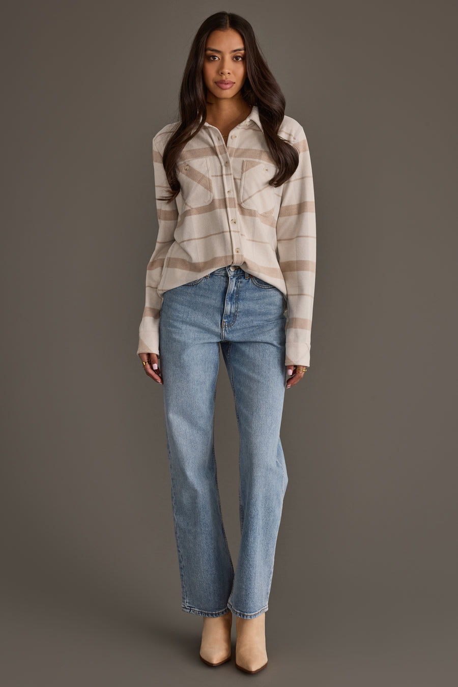 A woman stands against a gray background wearing the Cream, Tan & Gray Plaid Button Up from Panache Apparel Collection, paired with light blue jeans and tan heels. Her long hair is worn loose and she maintains a neutral expression, with her arms relaxed at her sides.