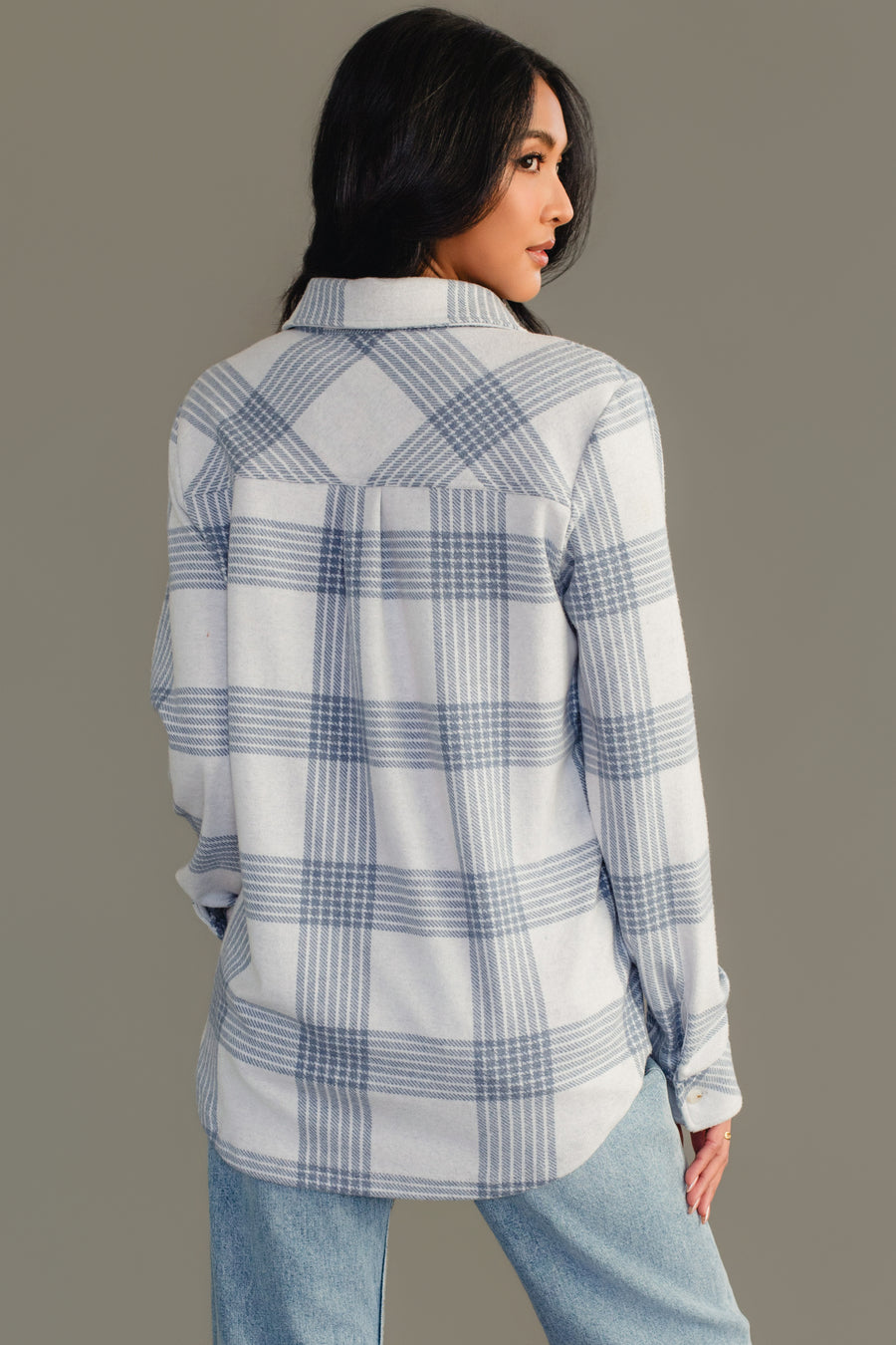 A woman with long dark hair wears the Panache Apparel Collection’s White & Dusty Blue Plaid Button Up, paired with light blue jeans, as she faces away and glances over her shoulder against a plain gray background.