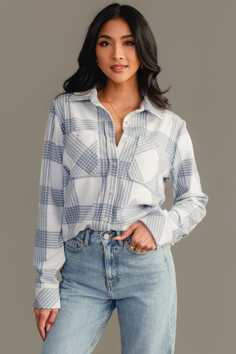 A woman with long dark hair is wearing the White & Dusty Blue Plaid Button Up from Panache Apparel Collection and light blue jeans, standing against a plain gray background with one hand in her pocket.