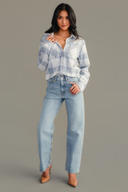 A woman stands against a plain gray background with one leg slightly forward, wearing the Panache Apparel Collection’s White & Dusty Blue Plaid Button Up paired with light blue jeans and tan high heels. She has long dark hair.