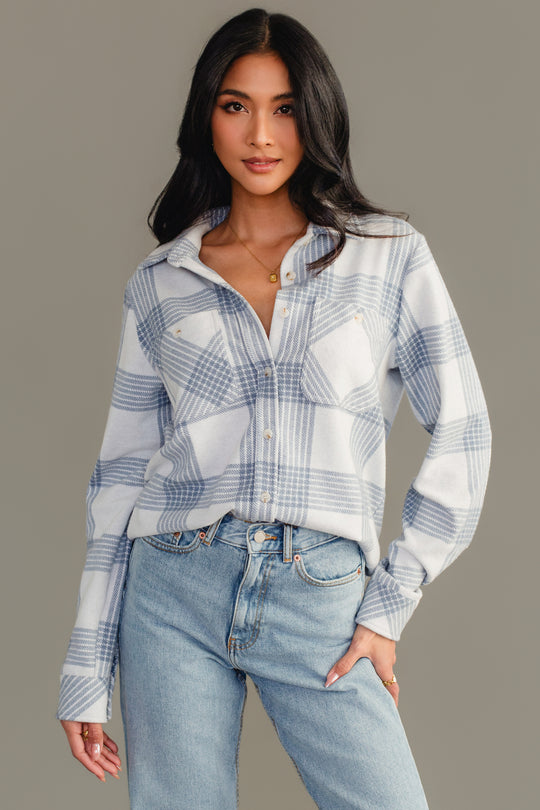 A woman with long dark hair is wearing the Panache Apparel Collection’s White & Dusty Blue Plaid Button Up paired with light blue jeans, standing against a plain gray background.