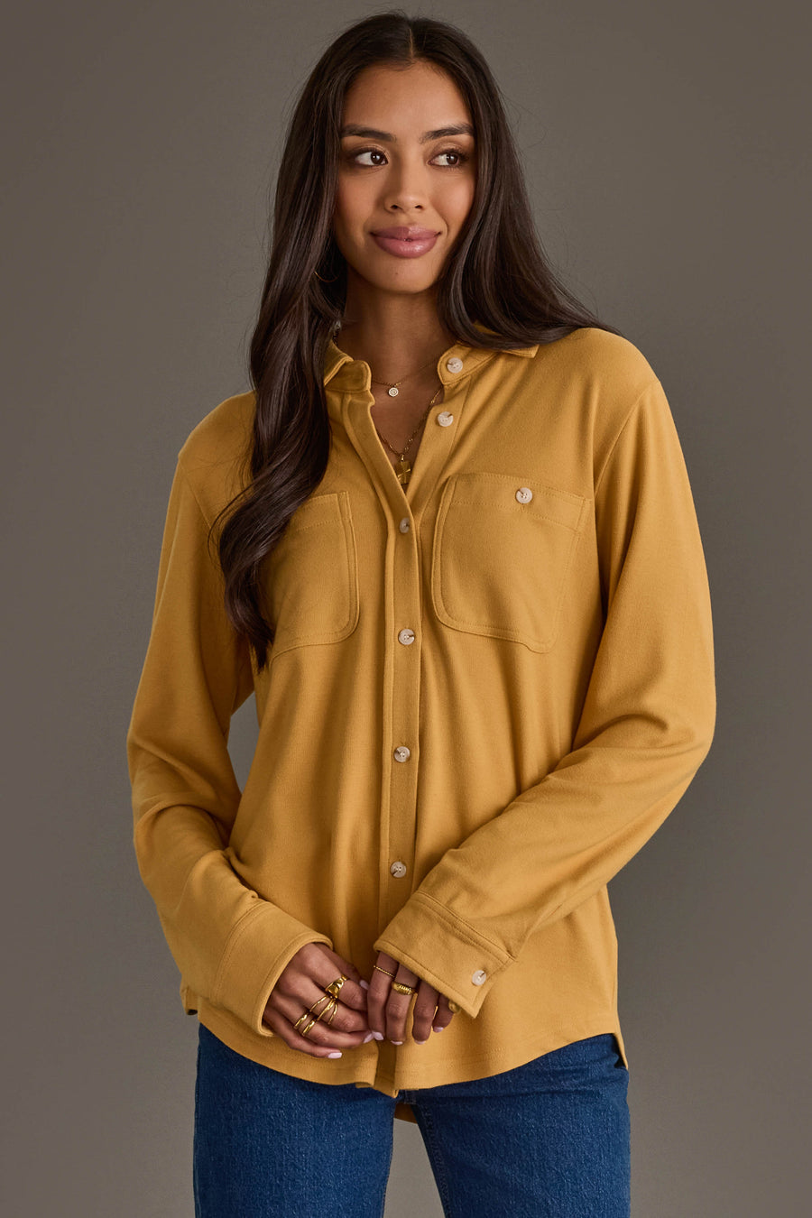 A woman with long dark hair wears the Mustard Super Soft Button Up from Panache Apparel Collection paired with blue jeans, standing against a plain gray background and looking slightly to the side with a soft smile.