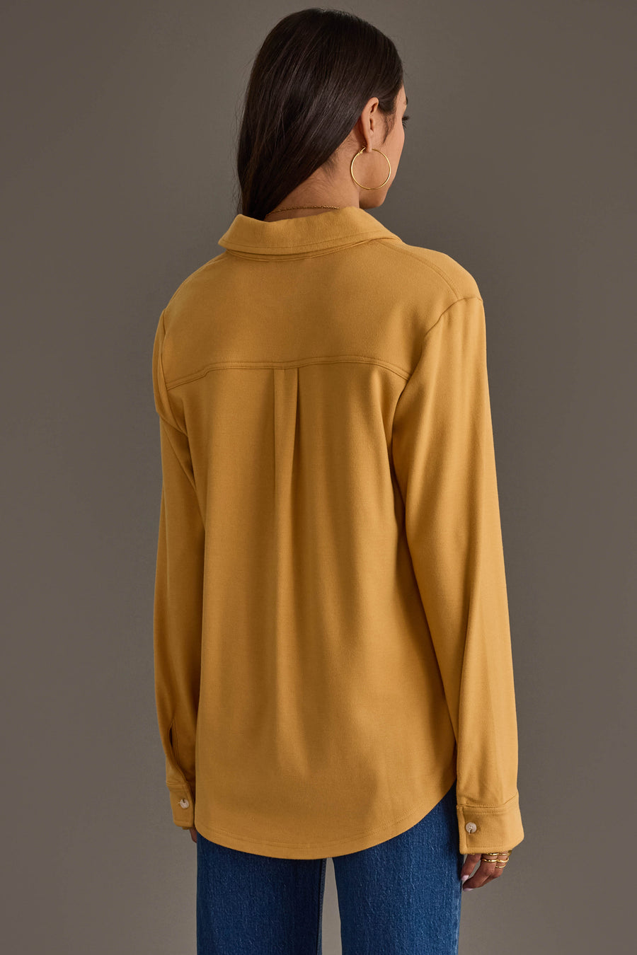 A woman with long brown hair, wearing gold hoop earrings, models the Panache Apparel Collection’s Mustard Super Soft Button Up with blue jeans, standing with her back to the camera against a plain brown background.