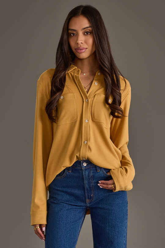 A woman with long dark hair stands against a plain gray background, looking at the camera with a slight smile and one hand in her pocket. She is wearing the Panache Apparel Collection Mustard Super Soft Button Up shirt paired with blue jeans.