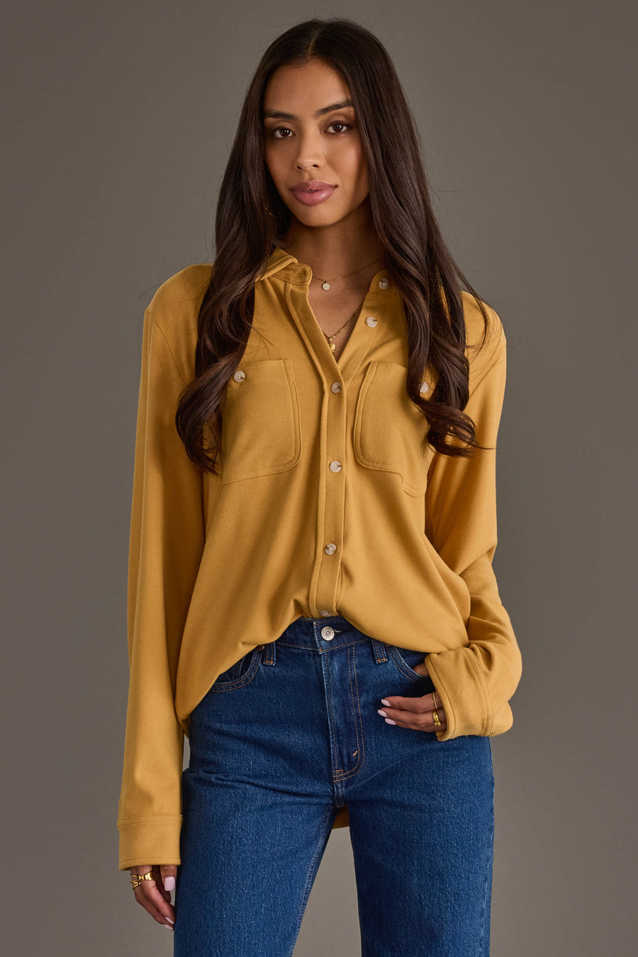 A woman with long dark hair stands against a plain gray background, looking at the camera with a slight smile and one hand in her pocket. She is wearing the Panache Apparel Collection Mustard Super Soft Button Up shirt paired with blue jeans.