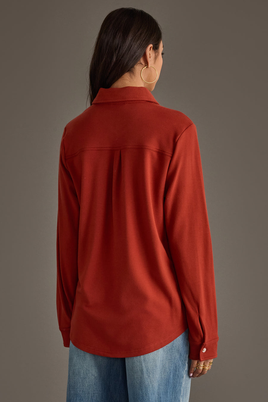 A woman with long dark hair, seen from behind against a plain brown background, models the Panache Apparel Collection’s Dark Rust Super Soft Button Up, paired with blue jeans.