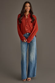 A woman with long dark hair is wearing the Panache Apparel Collection’s Dark Rust Super Soft Button Up, paired with wide-leg blue jeans, as she stands and smiles against a plain gray background.