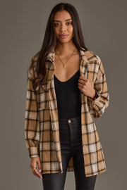 A woman stands facing forward against a plain background, wearing the Panache Apparel Collection’s Tan, Black & White Boyfriend Fit Plaid Button Up over a black top and black pants. Her long hair is down and she holds her shirt collar with one hand.