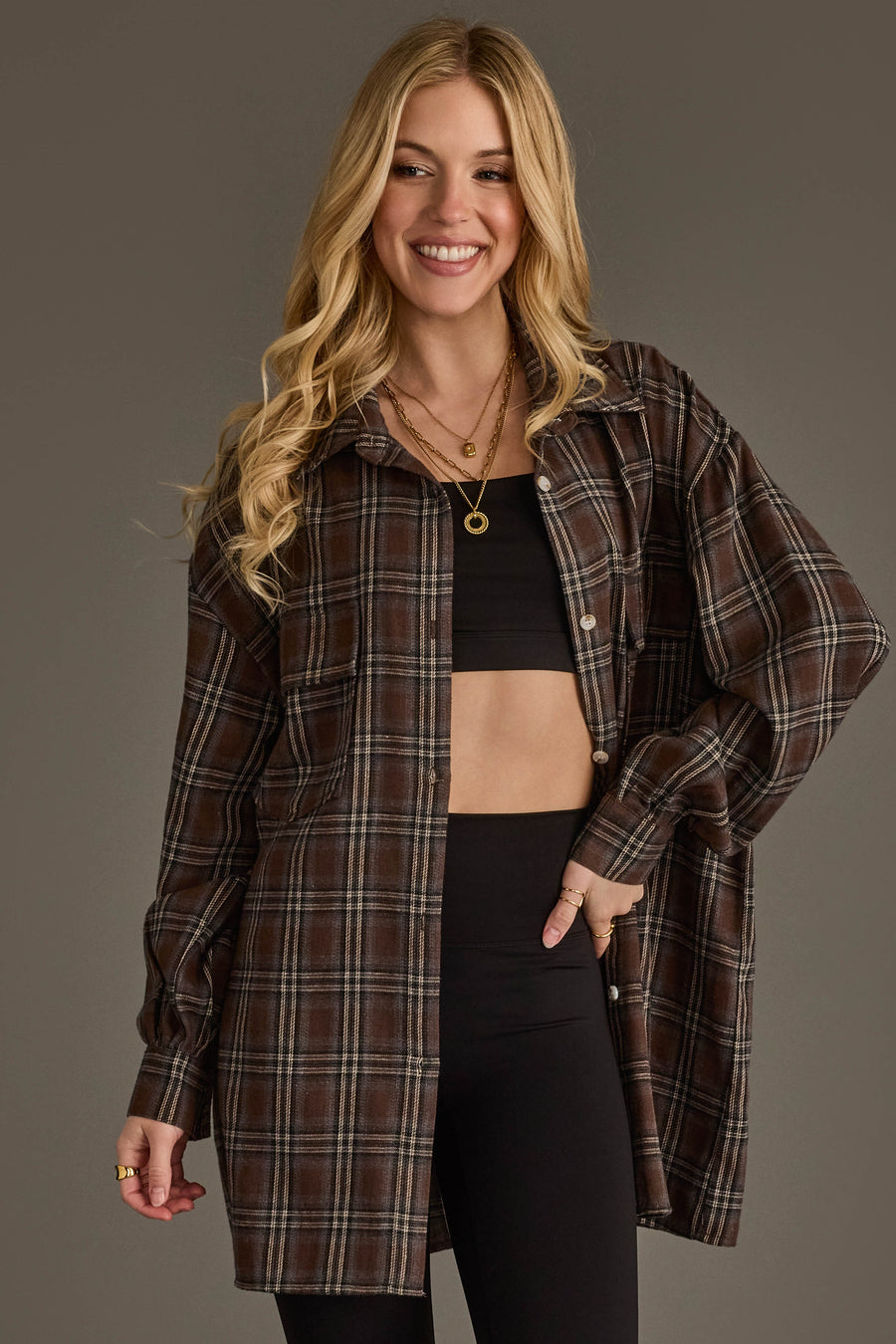 A woman with long blonde hair smiles, wearing the Panache Apparel Collection Brown, Blue, Cream & Black Boyfriend Fit Plaid Button Up over a black crop top and black pants, posing against a plain background.