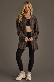 A woman with long blonde hair stands against a plain gray background, wearing the Panache Apparel Collection Brown, Blue, Cream & Black Boyfriend Fit Plaid Button Up over a black crop top and leggings, paired with white sneakers.