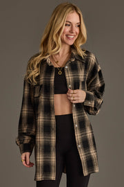 A smiling woman with long blond hair wears the Panache Apparel Collection Black & Tan Boyfriend Fit Plaid Button Up over a black crop top and black leggings, accessorized with gold necklaces and rings, standing against a plain gray background.