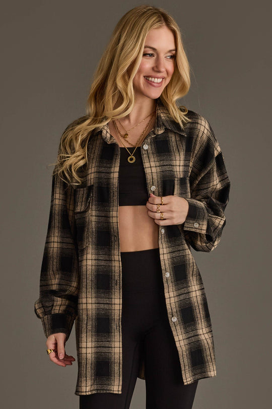 A smiling woman with long blond hair wears the Panache Apparel Collection Black & Tan Boyfriend Fit Plaid Button Up over a black crop top and black leggings, accessorized with gold necklaces and rings, standing against a plain gray background.