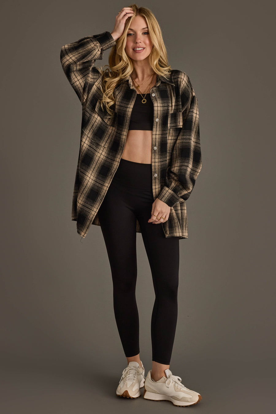 A woman wearing a black crop top, black leggings, white sneakers, and the Panache Apparel Collection Black & Tan Boyfriend Fit Plaid Button Up stands smiling with one hand on her head against a plain background.