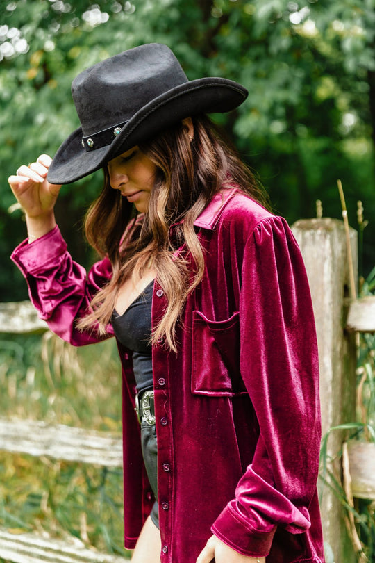 A person wearing a Panache Apparel Collection Burgundy Velvet Button Up and a black cowboy hat standing by a wooden fence.