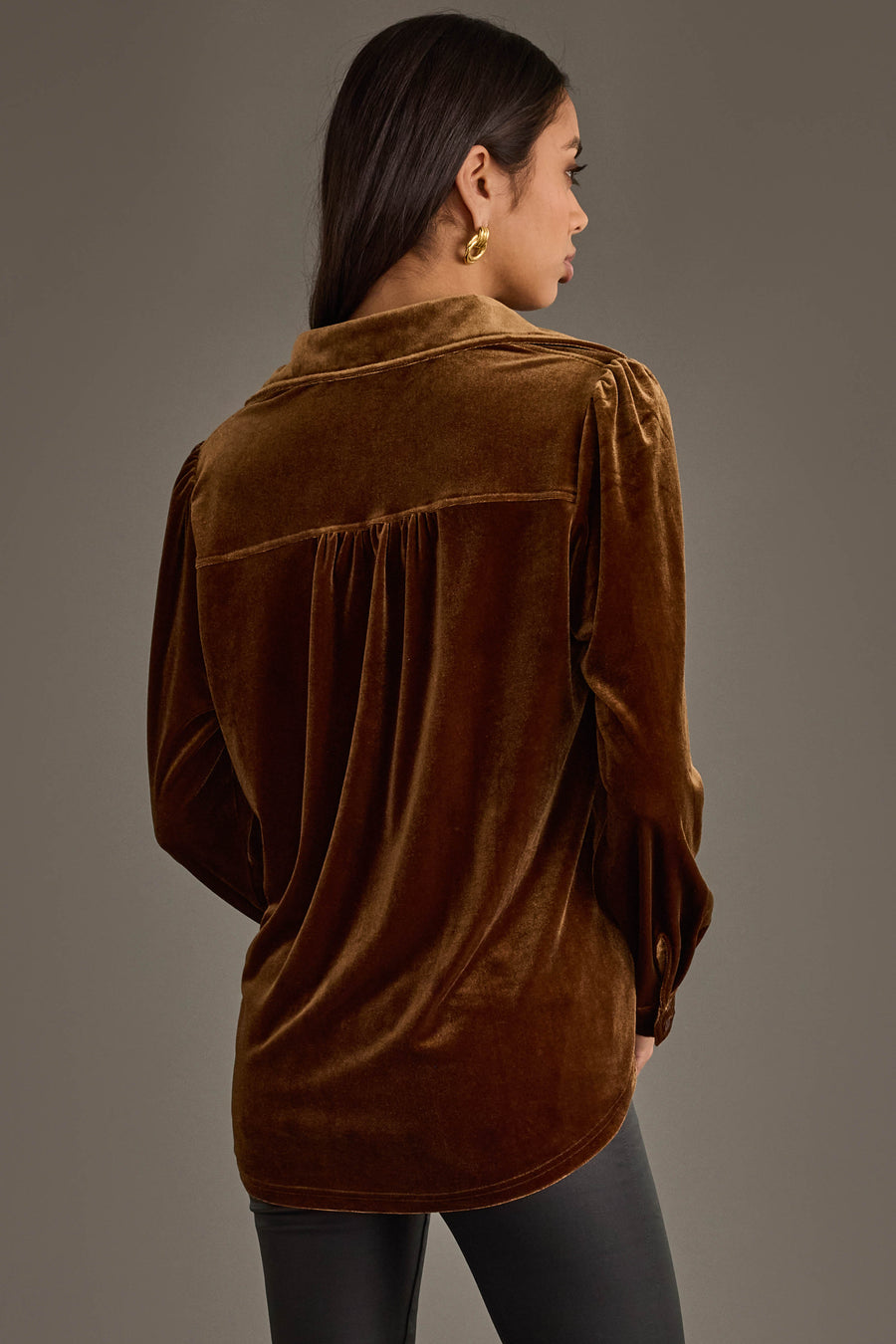 Woman with long dark hair wears the Bronze Velvet Button Up by Panache Apparel Collection and gold hoop earrings, shown from behind against a plain gray background. She pairs it with dark pants.