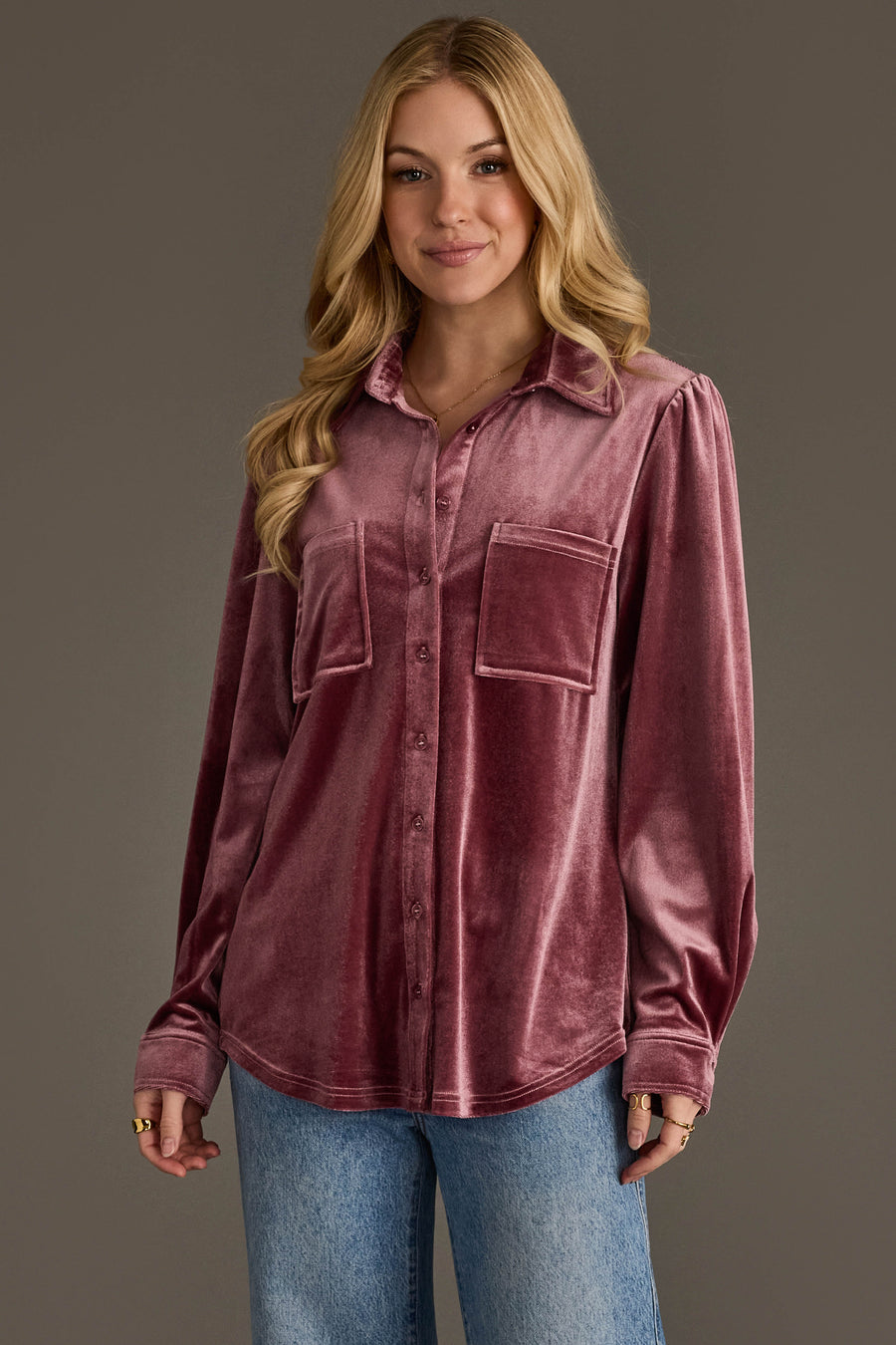 A woman with long blonde hair smiles at the camera, wearing the Mauve Velvet Button Up from Panache Apparel Collection with blue jeans, standing against a plain gray background.