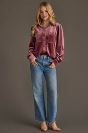 A woman with long blonde hair wears the Mauve Velvet Button Up by Panache Apparel Collection, tucked into high-waisted blue jeans and paired with tan heels, smiling slightly against a plain brown background.