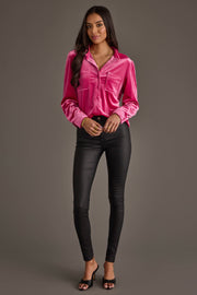 A woman poses against a gray background wearing the Bright Pink Velvet Button Up from Panache Apparel Collection, paired with black skinny pants and black open-toe heels. She has long dark hair and minimal jewelry.