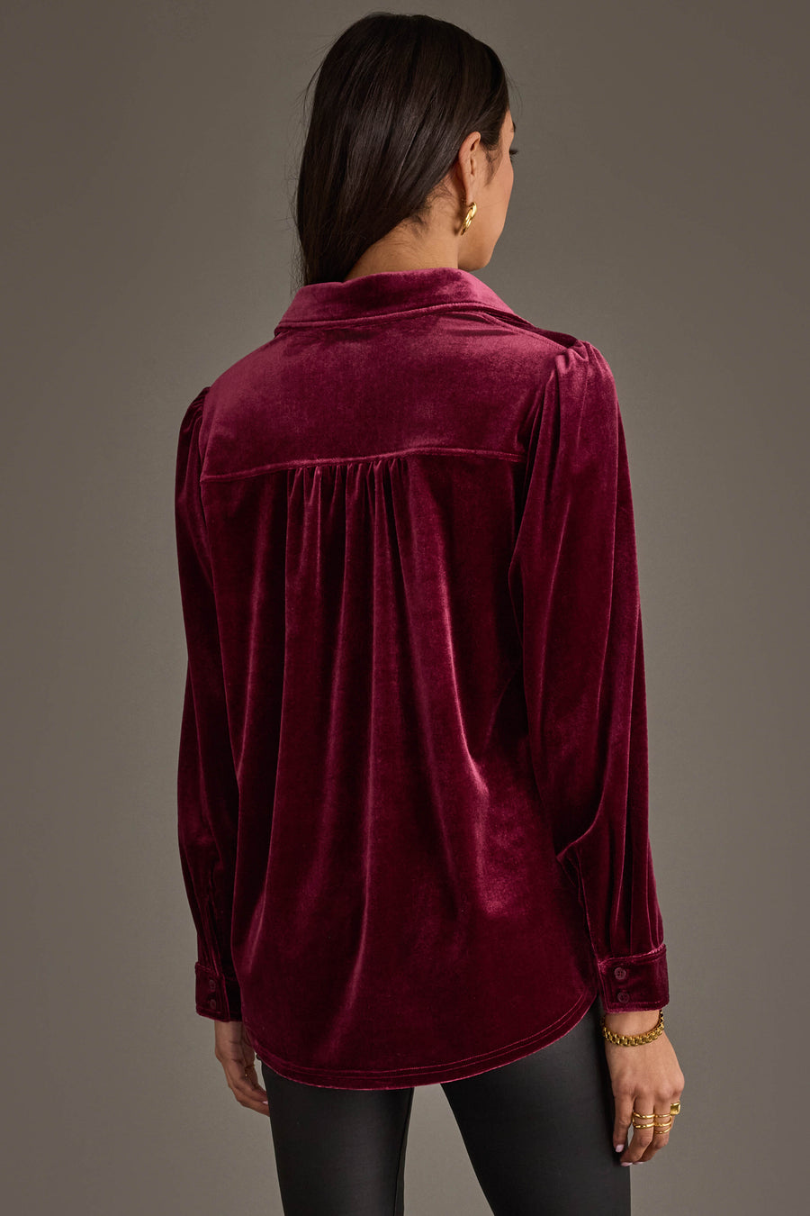 A woman with dark hair faces away, wearing the Maroon Velvet Button Up by Panache Apparel Collection with black pants, standing against a plain gray background.