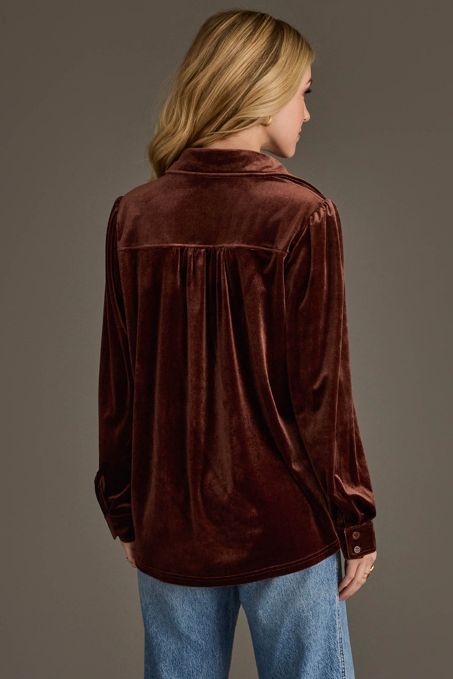 A woman with blonde hair, seen from behind, wears the Brown Velvet Button Up by Panache Apparel Collection with light blue jeans, standing against a dark background.