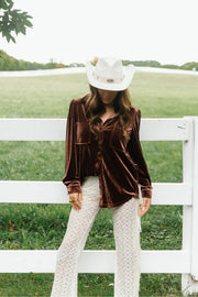 A woman wearing a Brown Velvet Button Up, white lace pants, and a white cowboy hat leans against a white wooden fence in a grassy field.