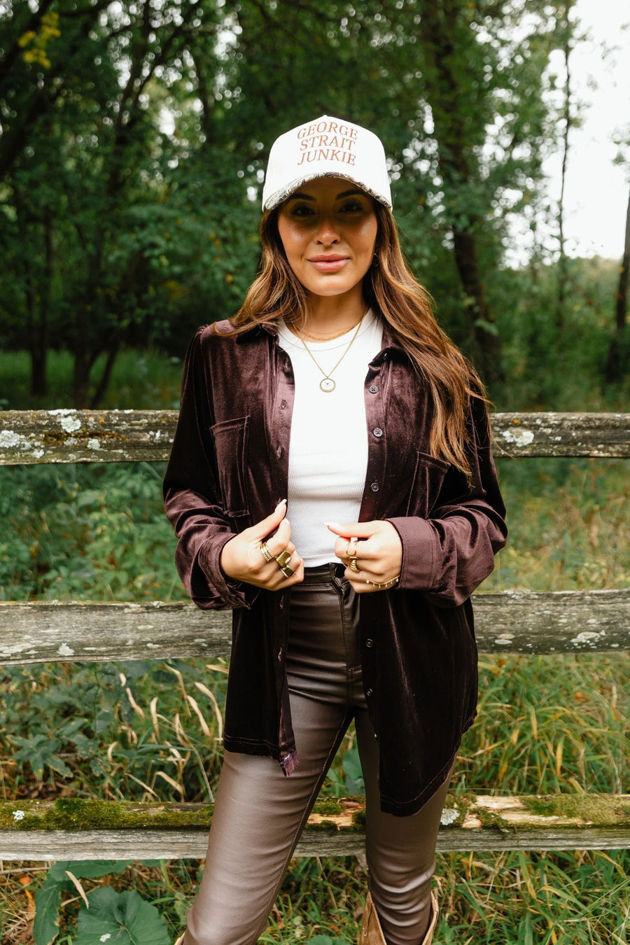 A person wearing a relaxed fit, espresso brown Espresso Brown Velvet Button Up over a white top, brown pants, and a text-adorned baseball cap.