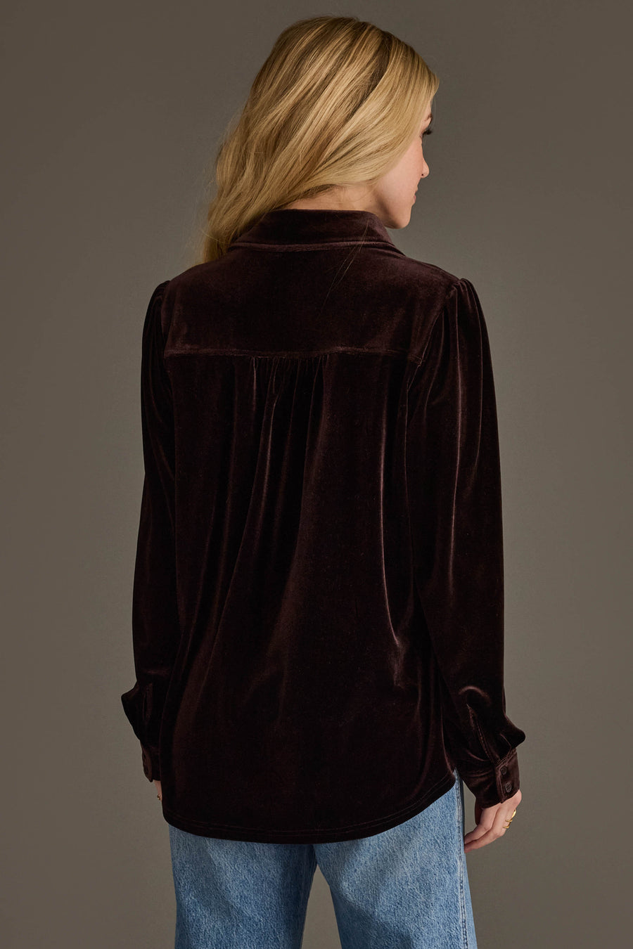 A woman with long blonde hair, seen from behind, wears the Espresso Brown Velvet Button Up from Panache Apparel Collection and light blue jeans against a plain gray background.