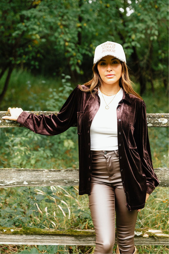 A person wearing an Espresso Brown Velvet Button Up over a white top and brown pants, with a cap that reads George Strait Junkie, leaning against a wooden fence.