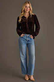 A woman stands against a gray background, wearing the Espresso Brown Velvet Button Up from Panache Apparel Collection with light blue high-waisted jeans and beige heels. She smiles slightly, hands in her pockets.