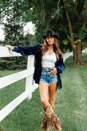 A woman wearing a relaxed fit, navy velvet Navy Velvet Button Up over a white crop top, denim shorts, and tan fringed boots, leaning against a white fence.