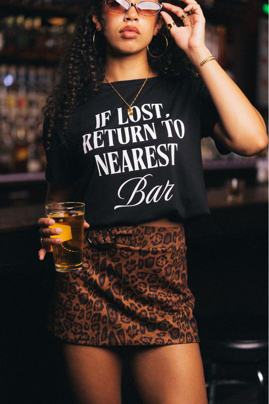 A woman in sunglasses holds a drink and stands in a lively bar, wearing the Panache Apparel Collection’s If Lost, Return To Nearest Bar Tee—a black graphic shirt—paired with a leopard print skirt.