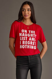 A person wearing a relaxed fit red On The Naughty List Tee that reads ON THE NAUGHTY LIST AND I REGRET NOTHING in white capital letters.