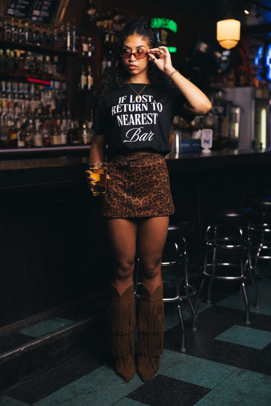 A woman in sunglasses, a leopard print skirt, and brown fringe boots stands in a bar holding a drink while wearing the Panache Apparel Collection’s “If Lost, Return To Nearest Bar Tee.”.